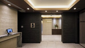 Un moderno vestíbulo de hotel con una mesa de recepción, una cabina de registro digital, un panel de control de ascensores en la pared y un arreglo floral sobre un pedestal.