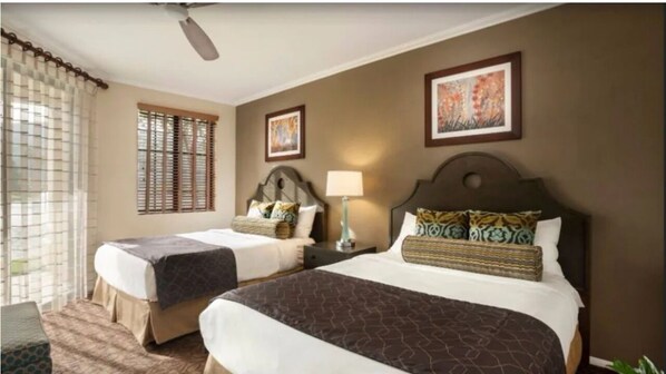 Habitación de hotel con dos camas, ventilador de techo, ventana con persianas y dos cuadros en la pared.