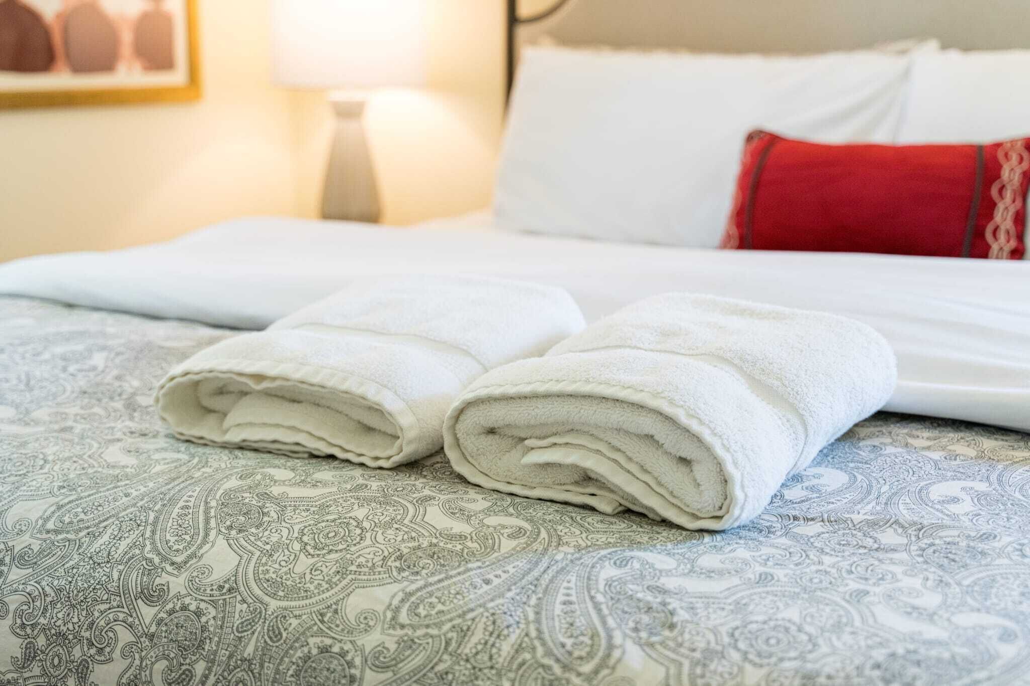 Een opgemaakt bed met witte lakens en een gevouwen handdoek op een beddekleed met dessin.