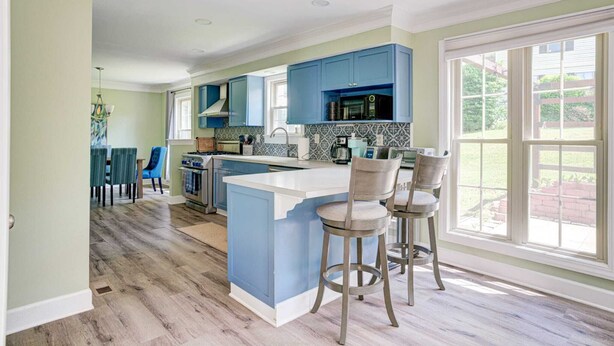 A kitchen with blue cabinets, a white island with bar stools, and a view of the backyard through large windows.