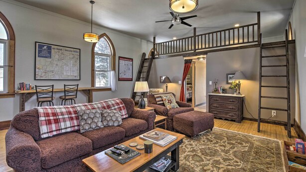 A spacious living room with a large sofa, a coffee table, and a staircase leading to an upper level. - Renovated Historic Church Along Namekagon River! (Hayward)