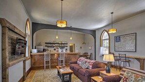 A cozy living room with a fireplace, wooden accents, a kitchen in the background, and a map on the wall. - Renovated Historic Church Along Namekagon River! (Hayward)
