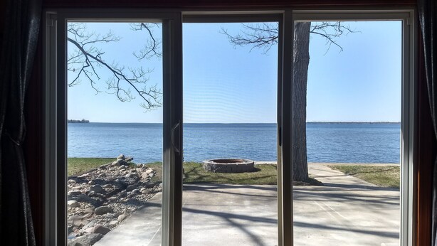 A room with a large sliding glass door leading to a view of a lake, a concrete path, and a stone structure.
