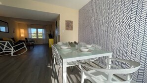 A dining area with a glass table and white chairs, a patterned wallpaper, and a view of the living room.