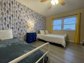A bedroom with two beds, a nightstand, a ceiling fan, and a window with yellow curtains.