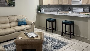 A living room with a beige sofa, a coffee table, and a rug. There is a kitchen area with wooden cabinets and a microwave.