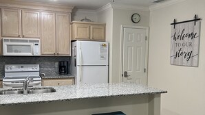 A kitchen with wooden cabinets, a white refrigerator, a microwave, a stove, and a sink. There are two stools and a welcome sign on the wall.