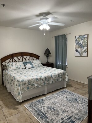 Ein Schlafzimmer mit Bett, Deckenventilator, Nachttisch, Lampe, Fenster mit Vorhängen und einem Bild an der Wand.