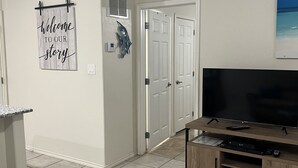 A living room with a flat-screen TV on a wooden stand, a 'Welcome to our story' sign on the wall, and a door leading to another room.
