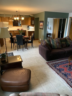 A living room with a sofa, ottoman, and a coffee table. There is a dining area with a table and chairs. The room has a kitchen in the background with wooden cabinets and a chandelier.