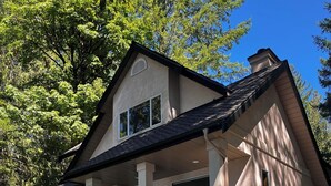 A two-story house with a covered patio, a grill, and a lounge chair. - Pet-Friendly Sproat Lake Suite | Private Dock, Paddleboard/Kayaks, Firepit (Port Alberni)