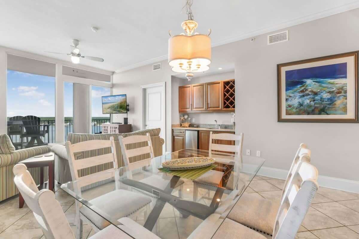 A spacious living area with a glass dining table, comfortable seating, a kitchen with wooden cabinets, and a large window offering a view.