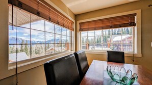 Ein Essbereich mit Tisch und Stühlen, große Fenster mit Holzjalousien und Blick auf Berge sowie eine Holzhütte.