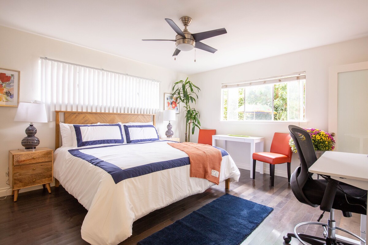 A bedroom with a bed, a desk, a chair, a ceiling fan, a window, and a plant.