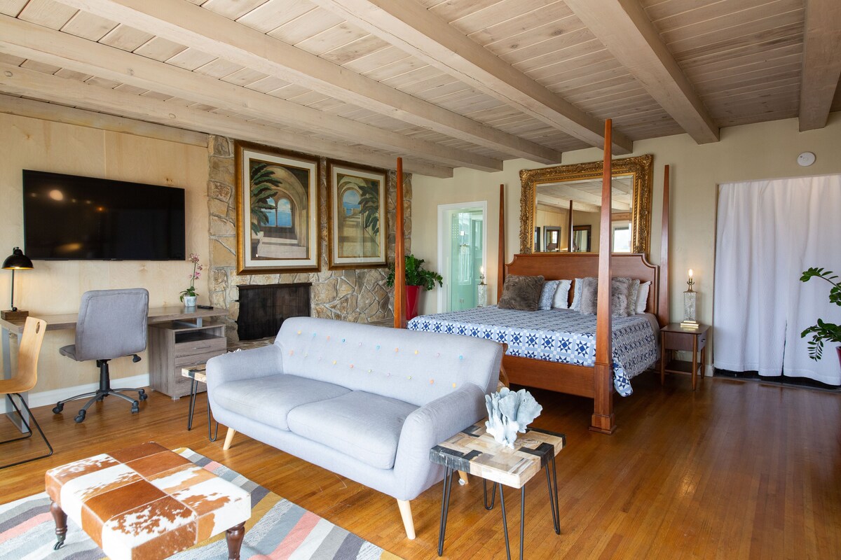 A spacious living room with a large sofa, a wooden coffee table, a TV mounted on the wall, a fireplace, and a four-poster bed.