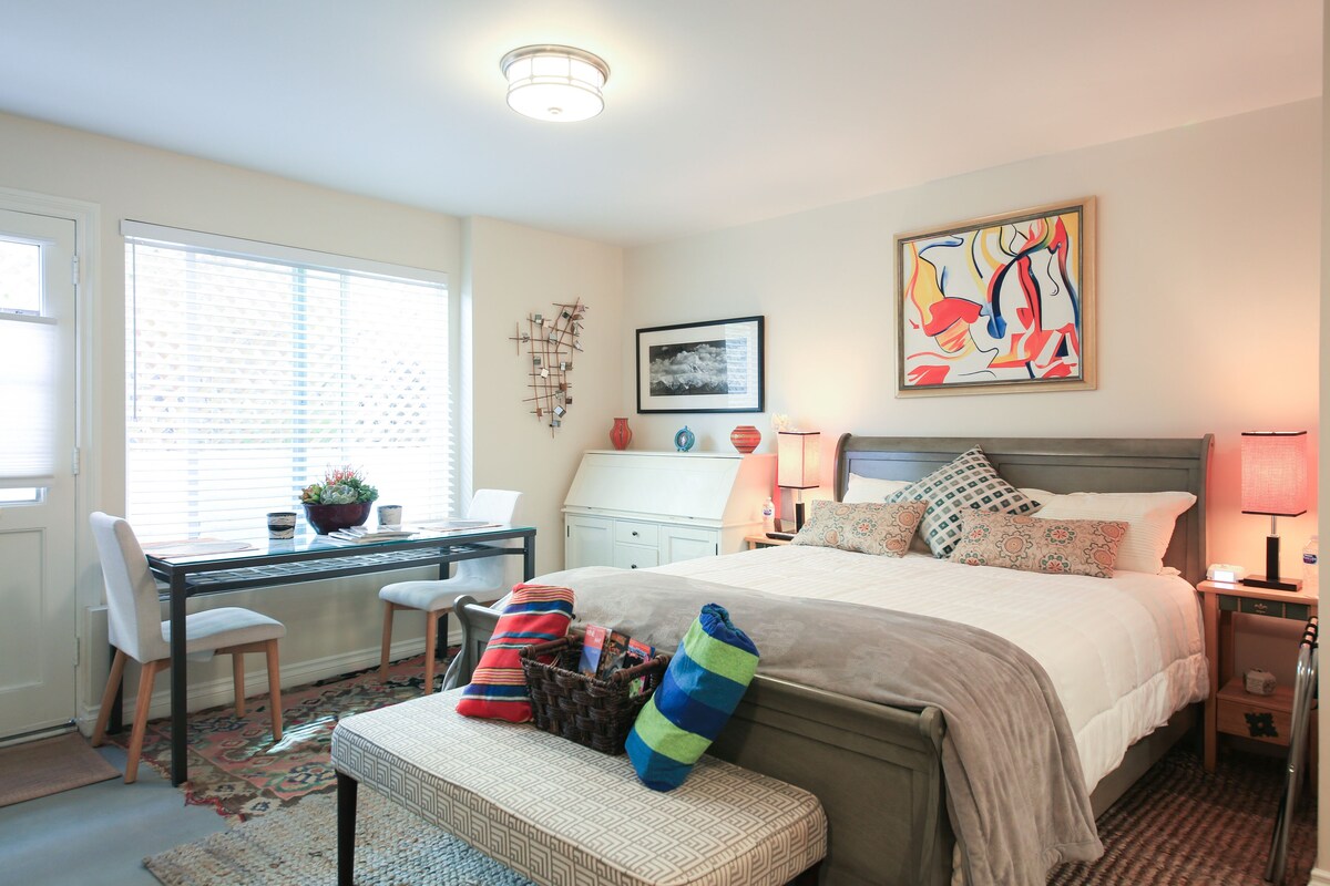 A bedroom with a bed, bedside tables, a desk with a chair, a window with blinds, and a wall with a framed picture.