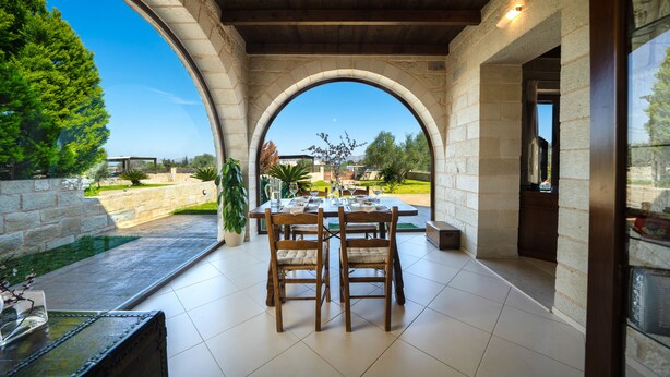 A covered patio with a table and chairs, stone walls, and a view of a garden.
