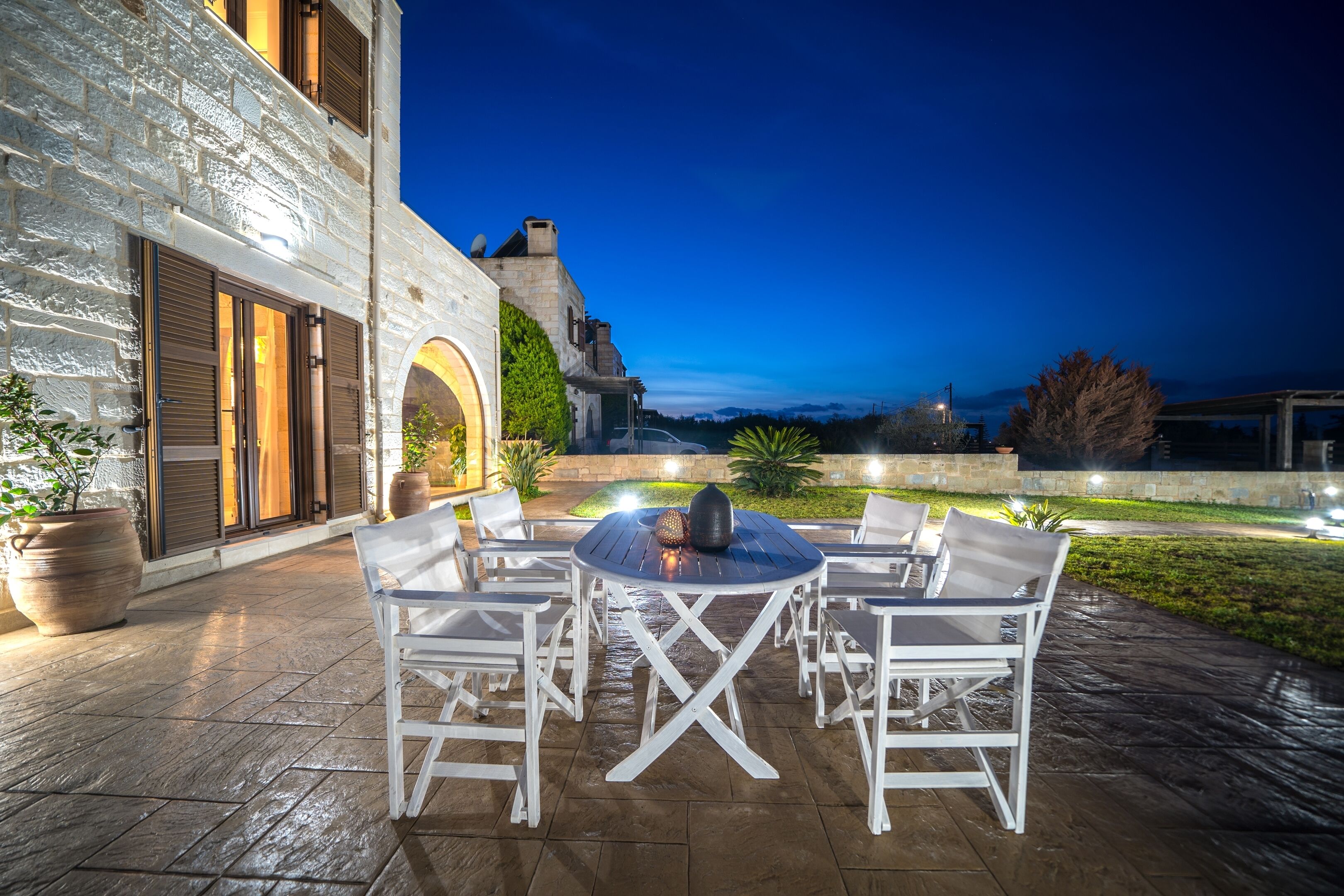 Une terrasse extérieure avec une table ronde et des chaises blanches, un bâtiment en pierre doté d’une grande porte en arc de cercle, et une vue sur le paysage au crépuscule.