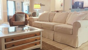 Un salon avec un canapé beige, une table basse en verre dotée de tiroirs en bois, un fauteuil à motifs, une lampe et, au fond, un coin cuisine.