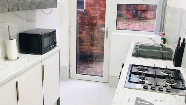 A compact kitchen with white cabinets, a black microwave, a gas stove, and a window overlooking a brick wall. - 4 Bedroom Lovely home in Loughborough town & Uni (Loughborough)