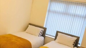 A room with two beds, each with a white mattress and a brown blanket. There is a window with blinds, and a light fixture on the ceiling. - 4 Bedroom Lovely home in Loughborough town & Uni (Loughborough)
