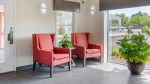 A hotel lobby with two red upholstered chairs, a potted plant, and a view of the parking area through large windows. - Rodeway Inn Mystic (Mystic)