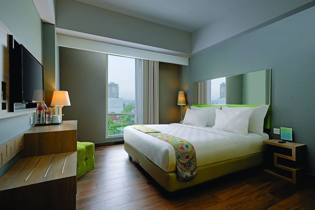 A modern hotel room with a large bed, a desk with a lamp, a TV, and a view of the city.
