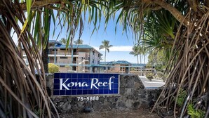 A sign for Kona Reef with a contact number, surrounded by tropical vegetation. - Private Oceanfront Top Floor Condo - Kona Reef D34 (Kailua-Kona)