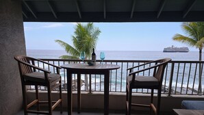 A balcony with two wooden chairs and a table, overlooking the sea with a ship in the distance. - Private Oceanfront Top Floor Condo - Kona Reef D34 (Kailua-Kona)