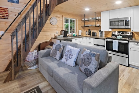 A cozy cabin interior with a staircase, grey sofa with patterned cushions, wooden walls, and a kitchen area with modern appliances.