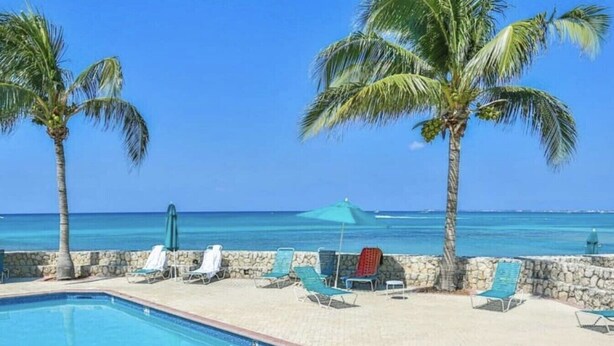 A pool area with lounge chairs and umbrellas, palm trees, and a view of the ocean. - 7Mile Pool/Seaside Bliss - 2bed 2bath Ground Suite (George Town)