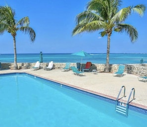 A pool area with lounge chairs and umbrellas, palm trees, and a view of the ocean.
