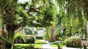A lush garden with a variety of plants and trees, a stone pathway, and a building in the background. - 7Mile Pool/Seaside Bliss - 2bed 2bath Ground Suite (George Town)