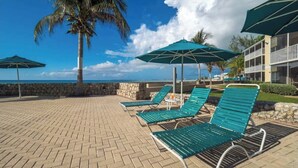 A beachside lounge area with green lounge chairs and umbrellas, a palm tree, and a view of the ocean. - 7Mile Pool/Seaside Bliss - 2bed 2bath Ground Suite (George Town)