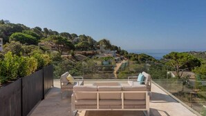 Un balcon avec un canapé et des fauteuils, donnant sur un paysage côtier avec des arbres et des maisons.