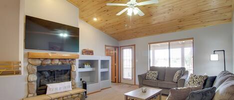 A living room with a stone fireplace, a flat-screen TV, a ceiling fan, and a sofa with patterned cushions.