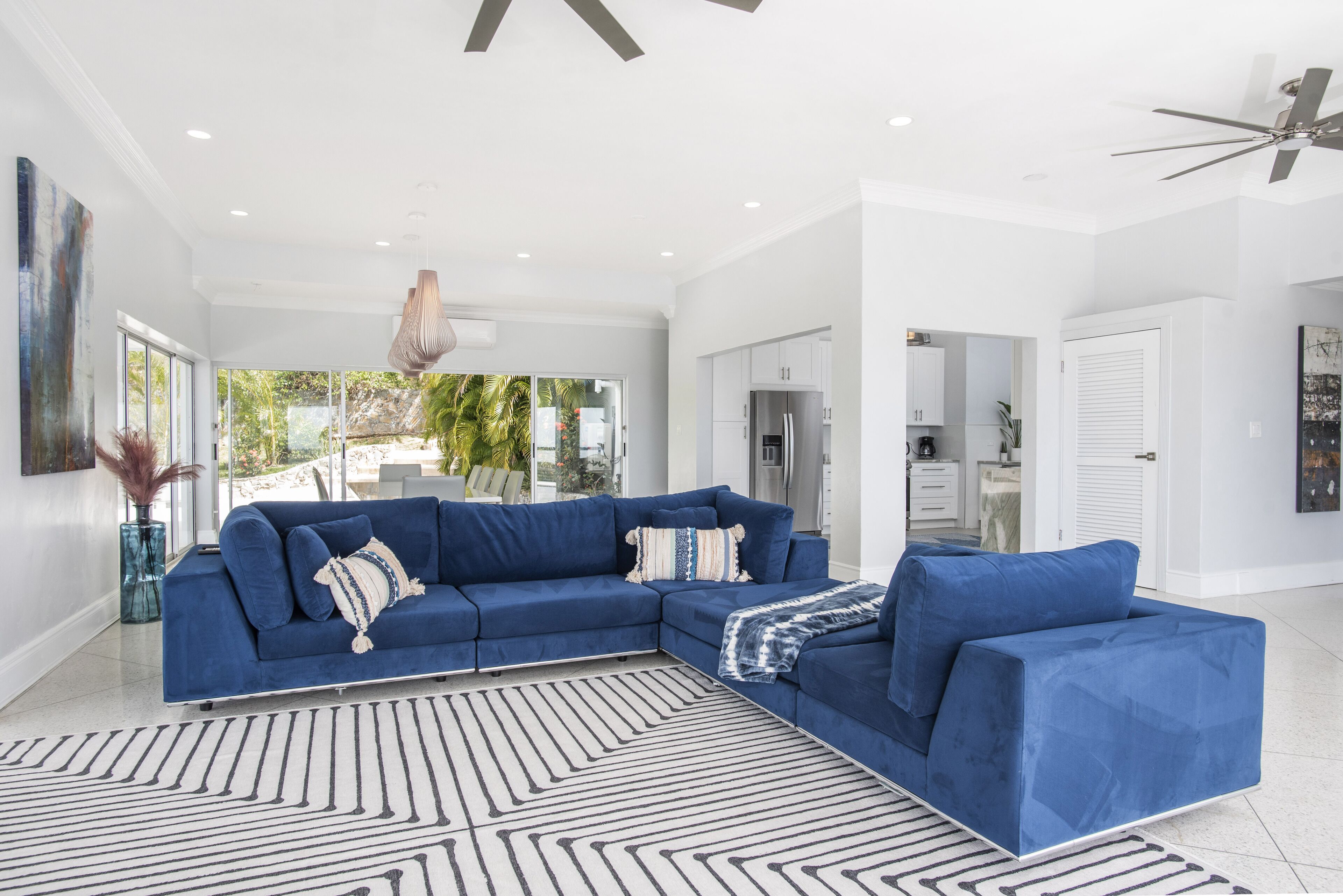 A modern living room with a blue sectional sofa, a patterned rug, and a view of the kitchen and outdoor area.