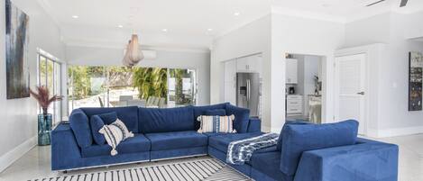 A modern living room with a blue sectional sofa, a patterned rug, and a view of the kitchen and outdoor area.