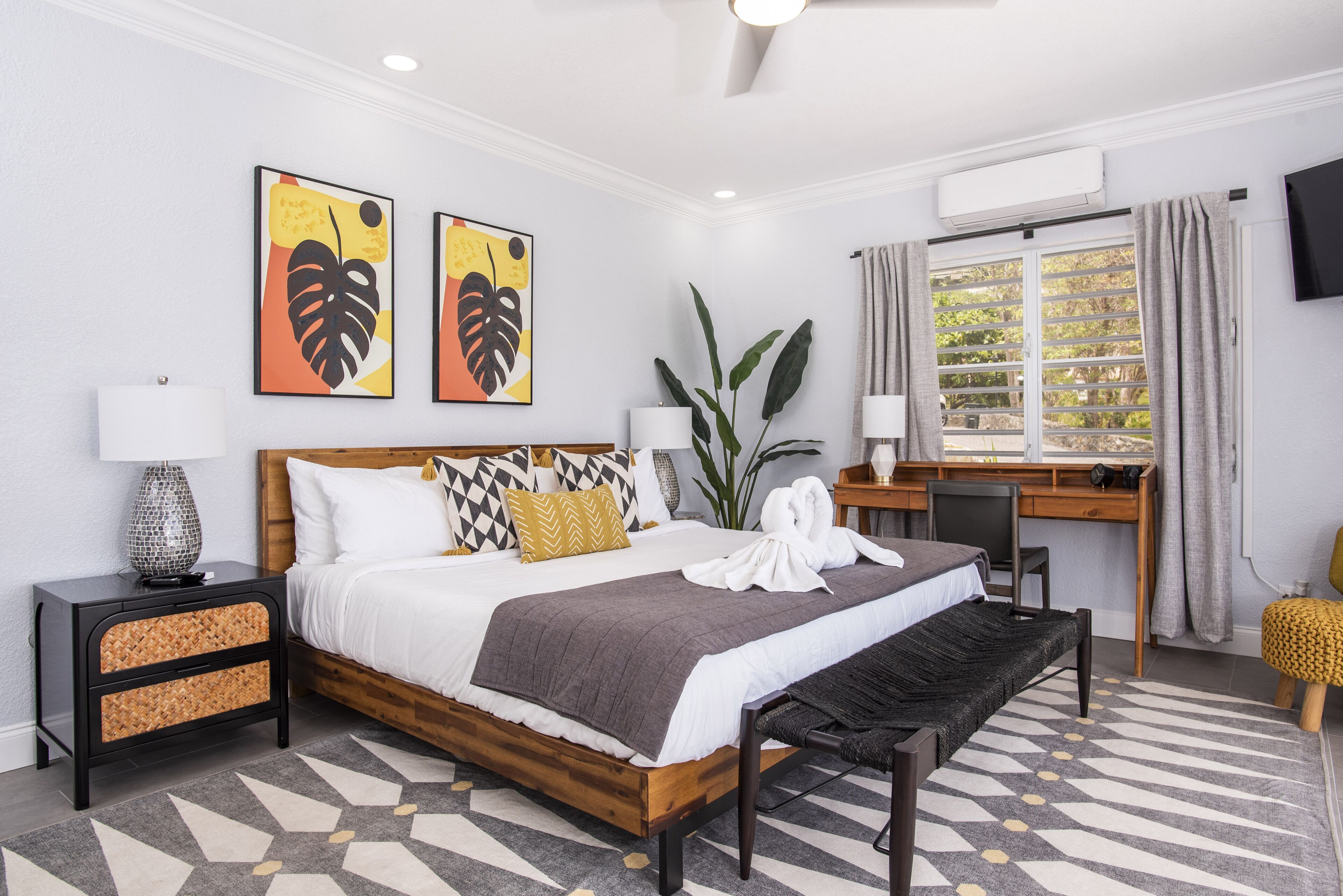 A hotel room with a wooden bed, a desk, a chair, a large window with curtains, and two wall-mounted artworks.