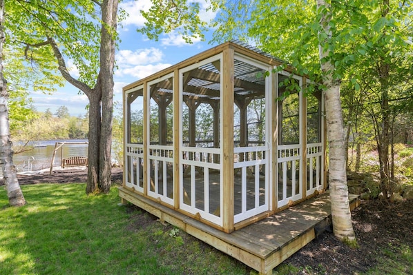 Ein hölzerner Pavillon mit Glasverkleidung, umgeben von Bäumen und einer Rasenfläche.