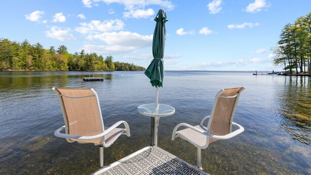 En vacker utsikt över en sjö med två stolar och ett bord under ett parasoll, omgivet av träd och en brygga i bakgrunden.