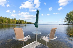 A clear view of a lake with two chairs and a table under an umbrella, surrounded by trees and a dock in the background.