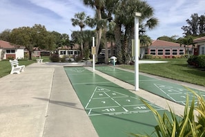 A shuffleboard court with green markings and a warning sign, surrounded by a grassy area with palm trees and a white bench.
