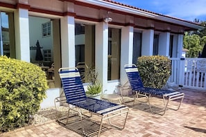 A covered outdoor seating area with two lounge chairs and a small table, surrounded by a white fence and greenery. - Walk to Beach & Fun! Heated Pool, Steps from Pool (Sarasota)