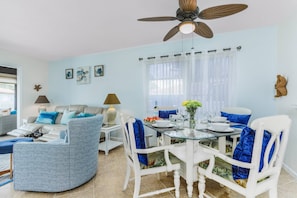 A modern living room with a glass dining table, white chairs, and a ceiling fan.