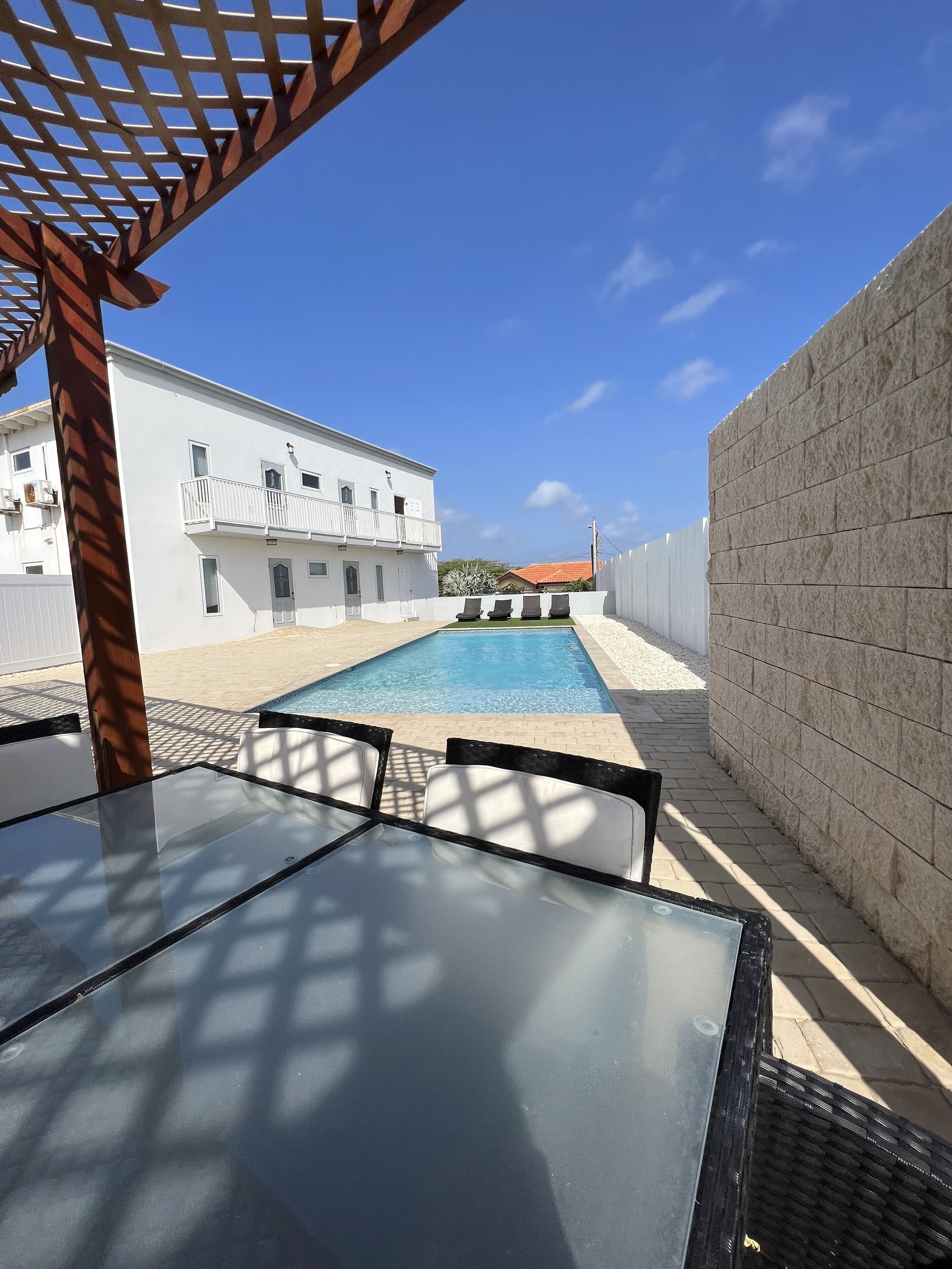 Zona de piscina con vista despejada a un edificio en el fondo, una estructura de pérgola y un camino pavimentado.
