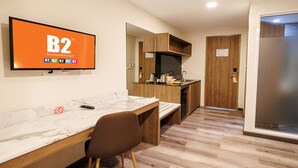 A hotel room with a TV displaying 'B2 Boutique&Budget', a wooden desk, a chair, a kitchen area, and a door. - B2 Khon Kaen Premier Hotel (Khon Kaen)