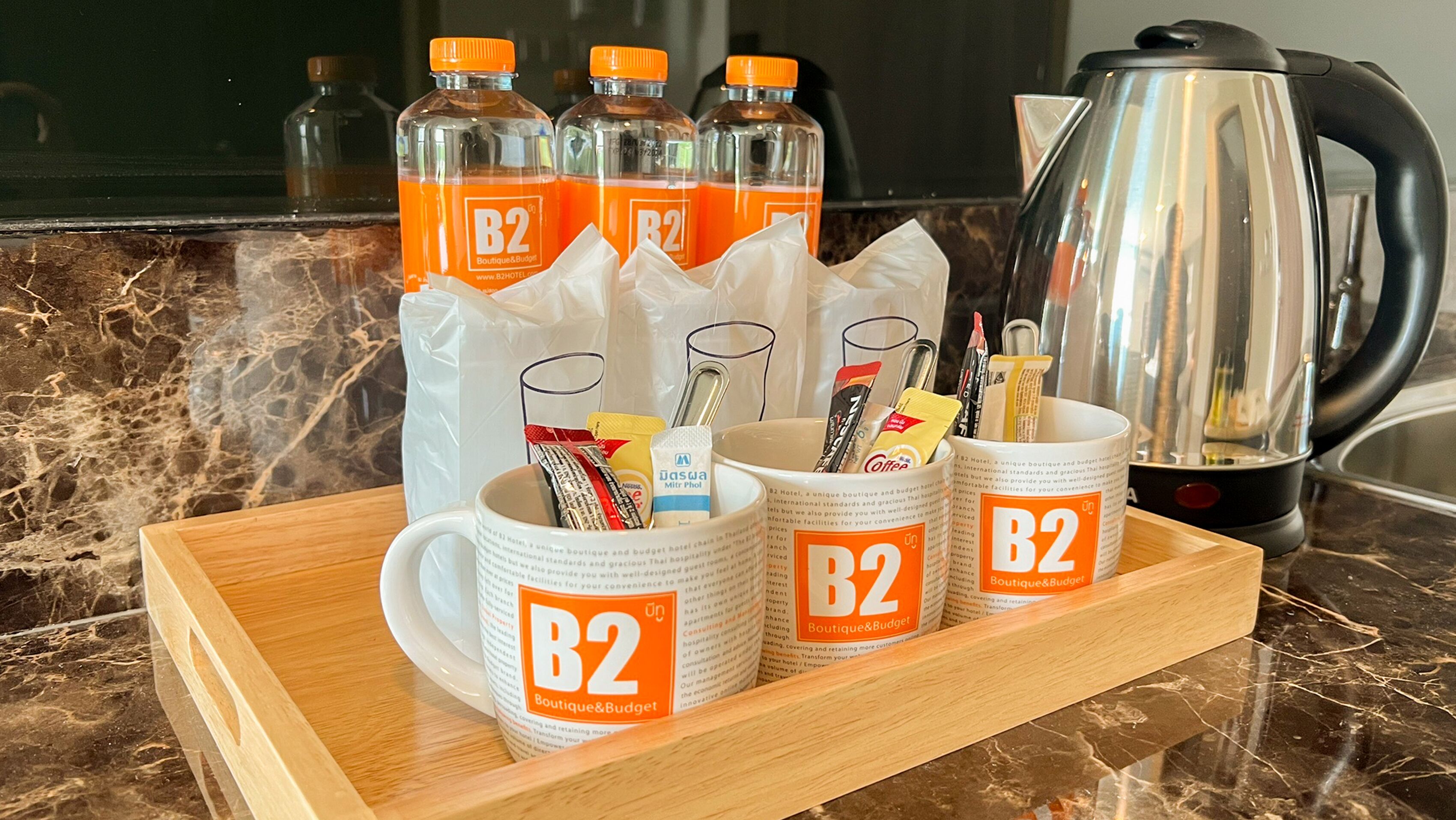 A tray with three B2 branded mugs, a coffee pot, and several bottles of orange juice on a marble countertop.