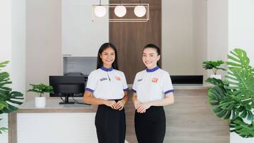 Two receptionists in white uniforms with blue collars standing in a modern hotel lobby with a wooden reception desk, a computer, and potted plants.