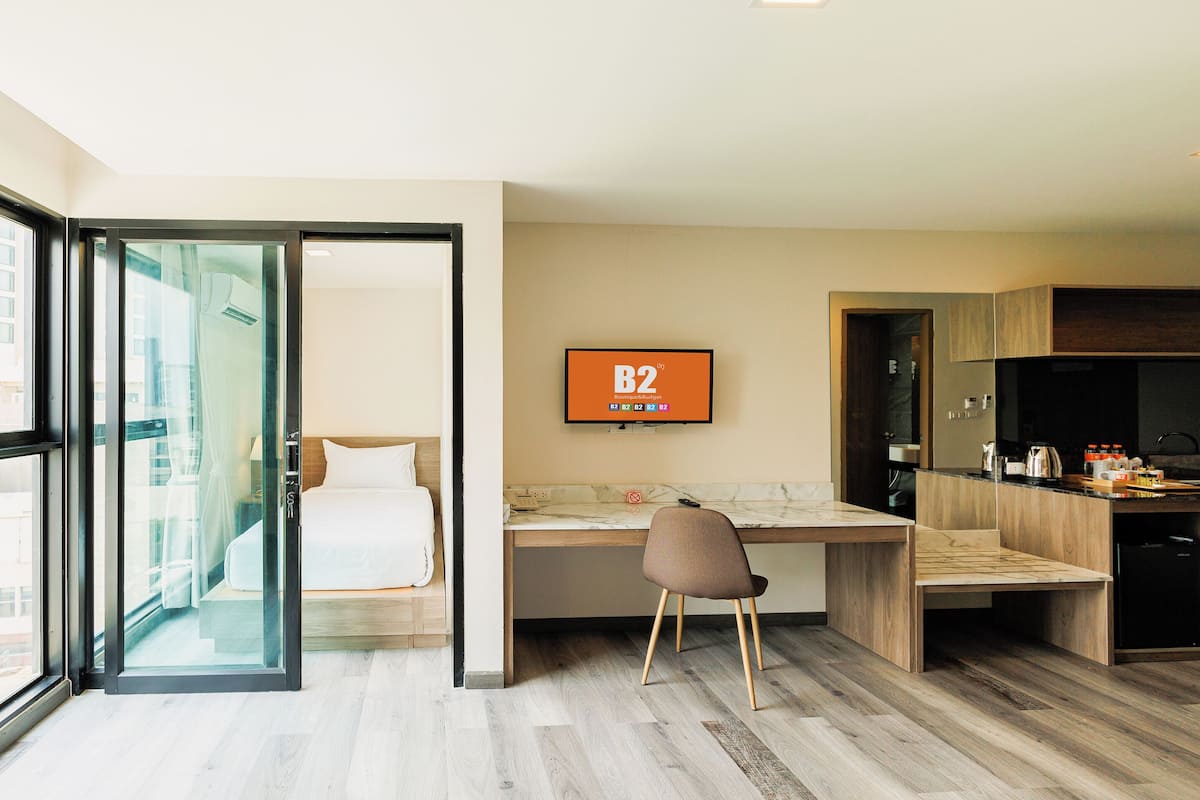 Une chambre d’hôtel moderne dotée d’une grande porte-fenêtre coulissante donnant sur un balcon, d’un lit, d’un bureau avec une chaise, d’un téléviseur fixé au mur et d’une kitchenette.